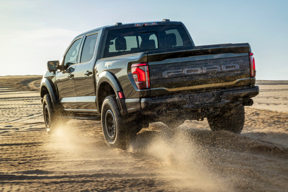 Rear view of a black Ford F-150 Raptor pickup truck driving on a sandy surface, kicking up dust.