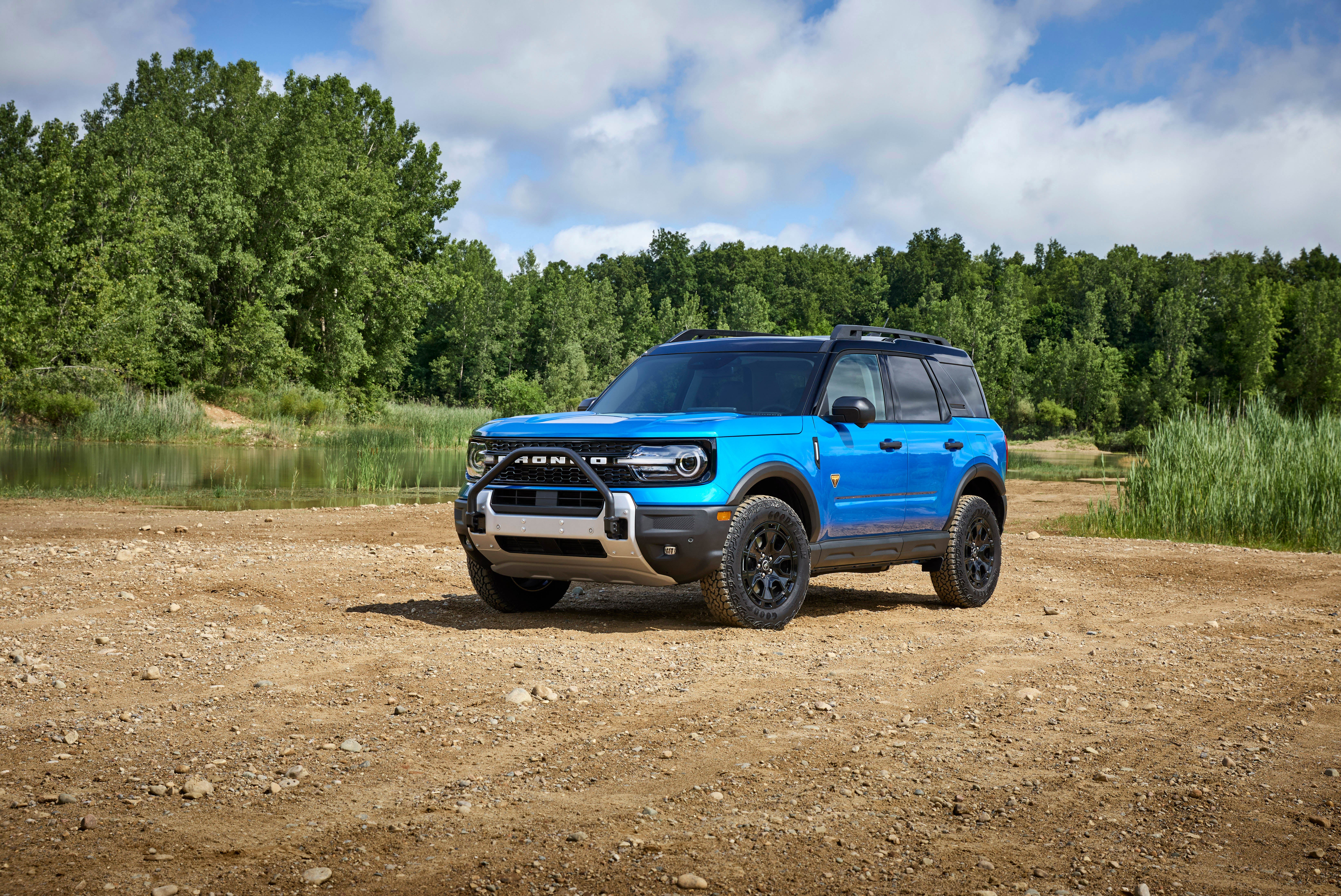 A medium shot of a blue Ford Bronco Sport SUV parked on a dirt path next to a body of water with a forest in the background.