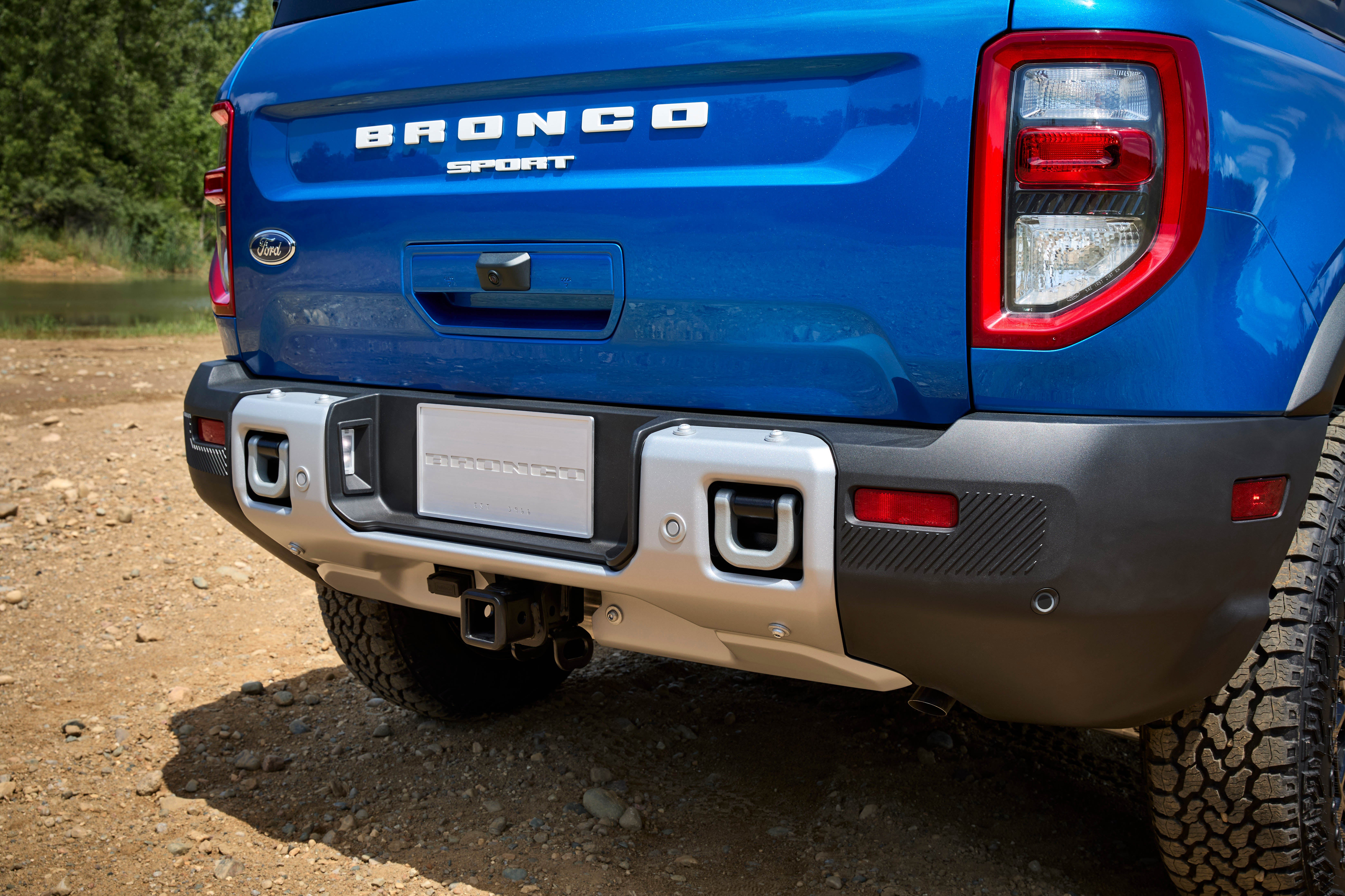 A close-up of the rear of a blue Ford Bronco Sport SUV, showing the 'BRONCO SPORT' lettering on the tailgate, the taillights, and the silver bumper with a hitch receiver and tow hooks. The vehicle is parked on a dirt path.