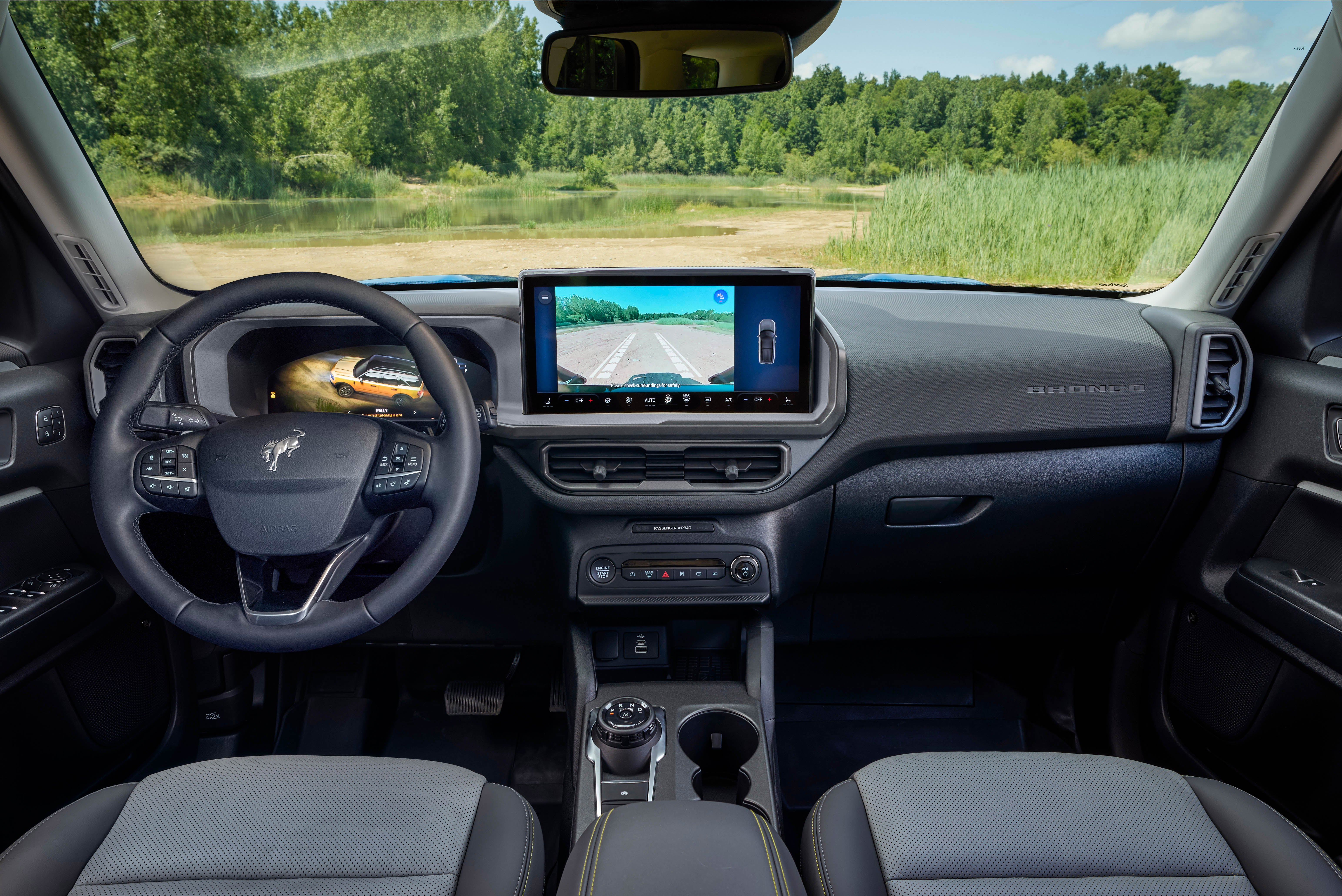 A full interior view from the driver's perspective of a blue Ford Bronco Sport SUV, showing the steering wheel, dashboard, digital instrument cluster, and a large center touchscreen displaying a rearview camera view. The car is parked next to a body of water with a forest in the background.