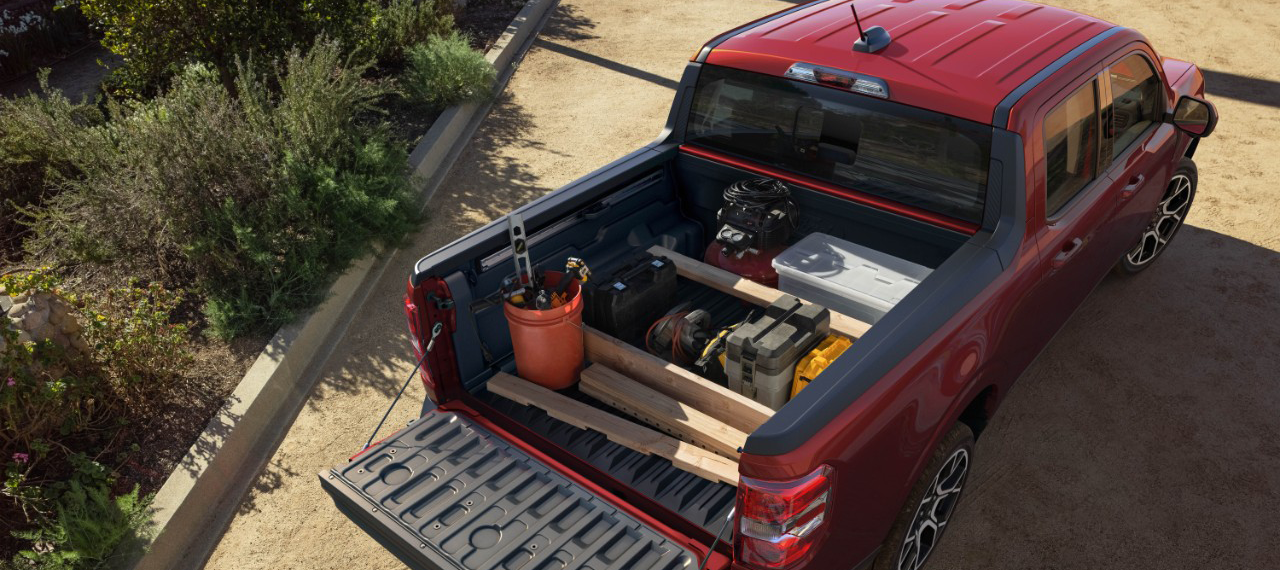 Red Ford Maverick pickup truck with its tailgate down, revealing a variety of items in the bed, including a toolbox, wood planks, and other equipment. 