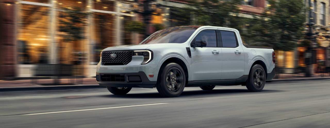 A white Ford Maverick compact pickup truck driving on a city street, with blurred buildings and trees in the background. 