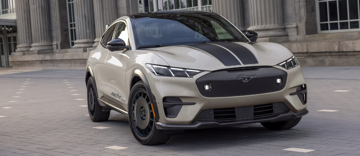 A light tan Ford Mustang Mach-E Rally edition, with black racing stripes and black wheels, parked on a paved area in front of a building with columns.