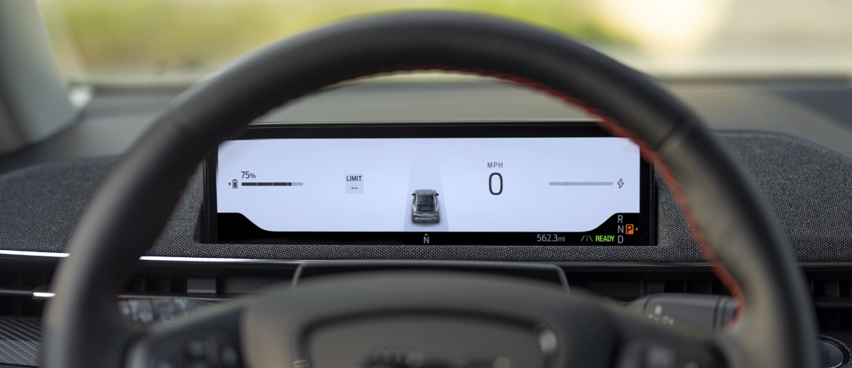A close-up view of a digital car dashboard seen through the steering wheel, showing a speedometer at 0 mph, a car icon, battery level, and mileage, with a red stitched detail visible on the steering wheel.
