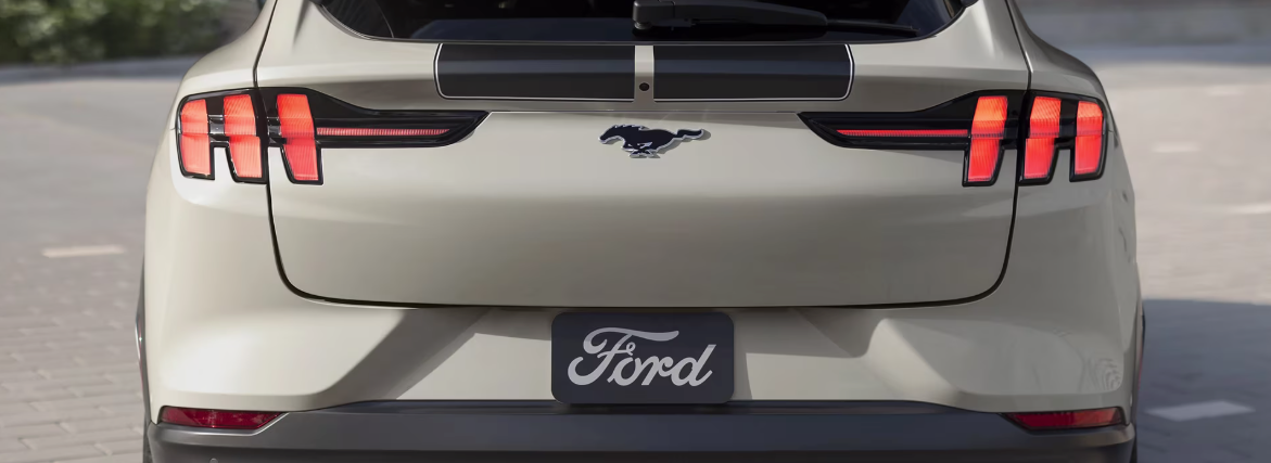 A rear view of a light tan Ford Mustang Mach-E, showing its distinctive three-bar LED taillights, a running horse emblem, black racing stripes, and a "Ford" badge on the license plate area.