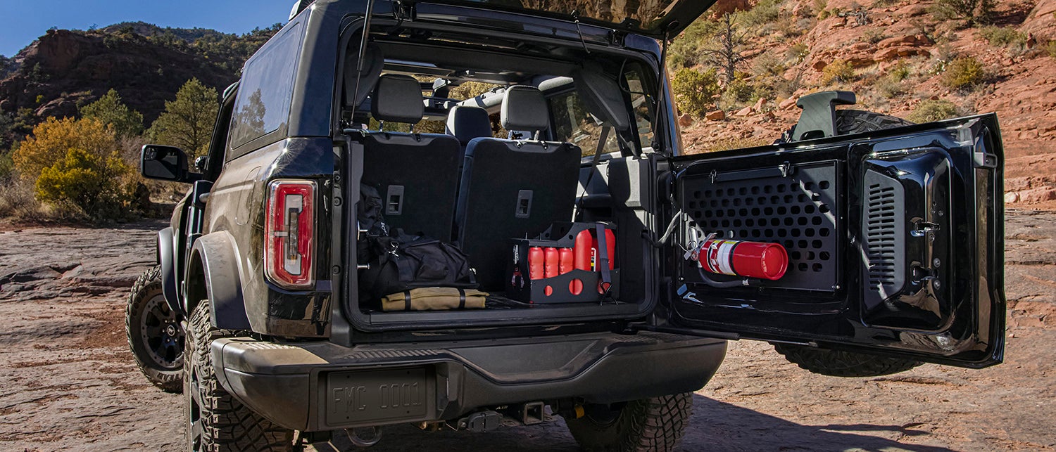 The interior of a Ford Bronco, showing a person in the driver's seat, the dashboard with a large touchscreen, and two-tone seats with a plaid pattern. The person is wearing a black shirt and has their hands on the steering wheel. Sunlight is streaming in through the windshield, illuminating the dashboard and the driver's side of the vehicle. The car is parked outdoors in a sunny location.