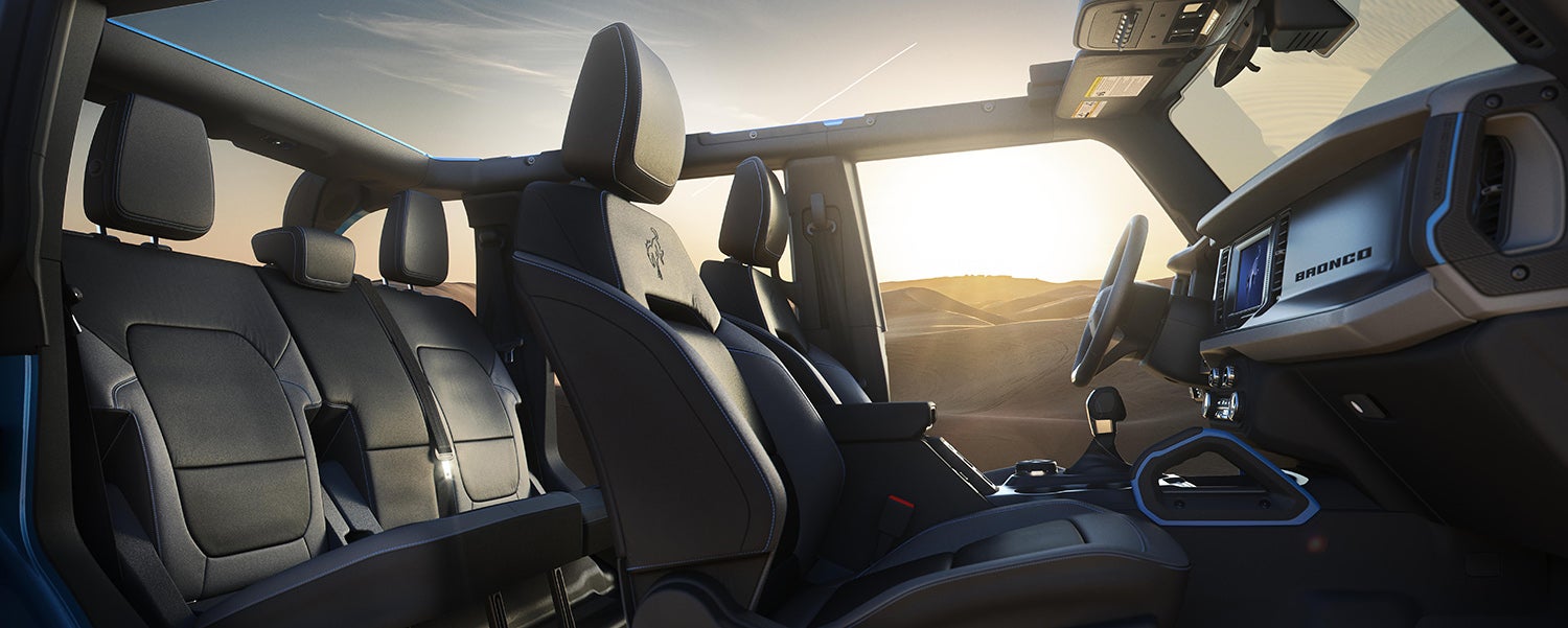 The interior of a Ford Bronco, showing a person in the driver's seat, the dashboard with a large touchscreen, and two-tone seats with a plaid pattern. The person is wearing a black shirt and has their hands on the steering wheel.