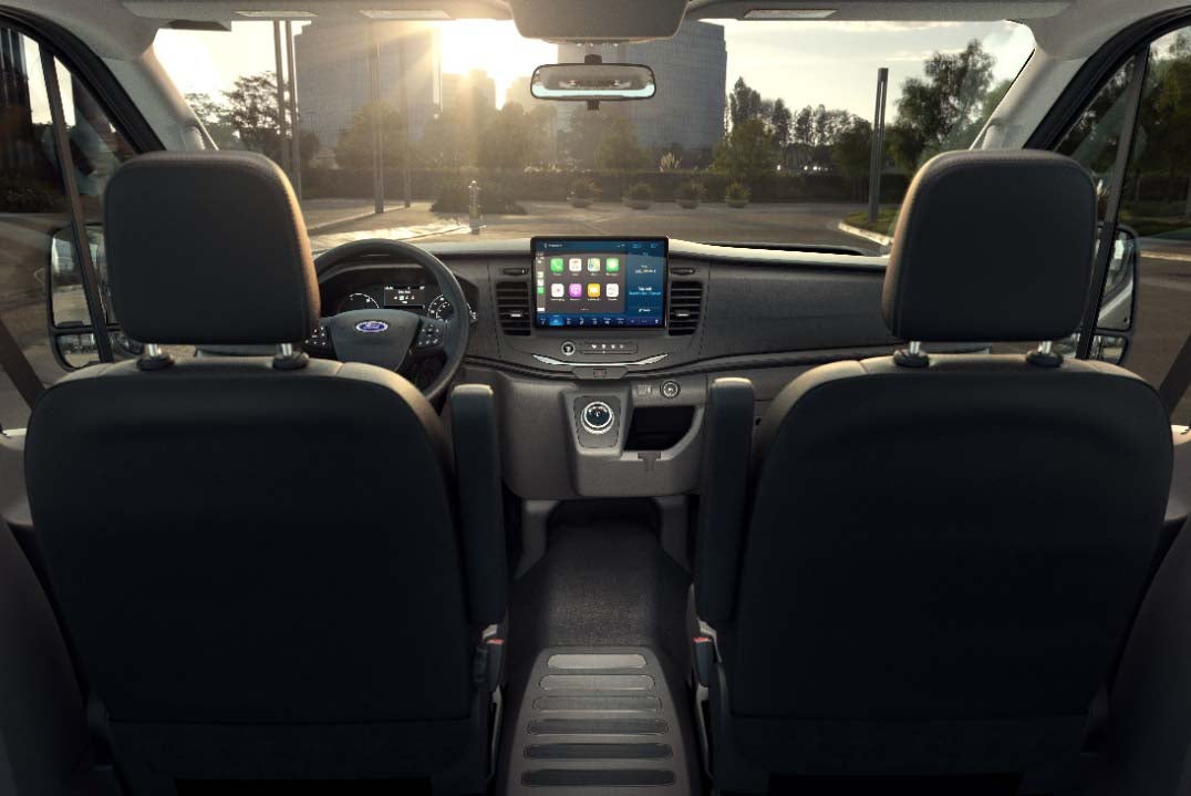 An interior shot of a Ford Transit van, taken from the back seats, looking forward towards the dashboard. The dashboard features a large, centrally-mounted touchscreen infotainment system displaying various app icons. The driver and passenger seats are visible in the foreground, and the steering wheel is on the left. The sun is setting in the background, visible through the windshield, with city buildings silhouetted.